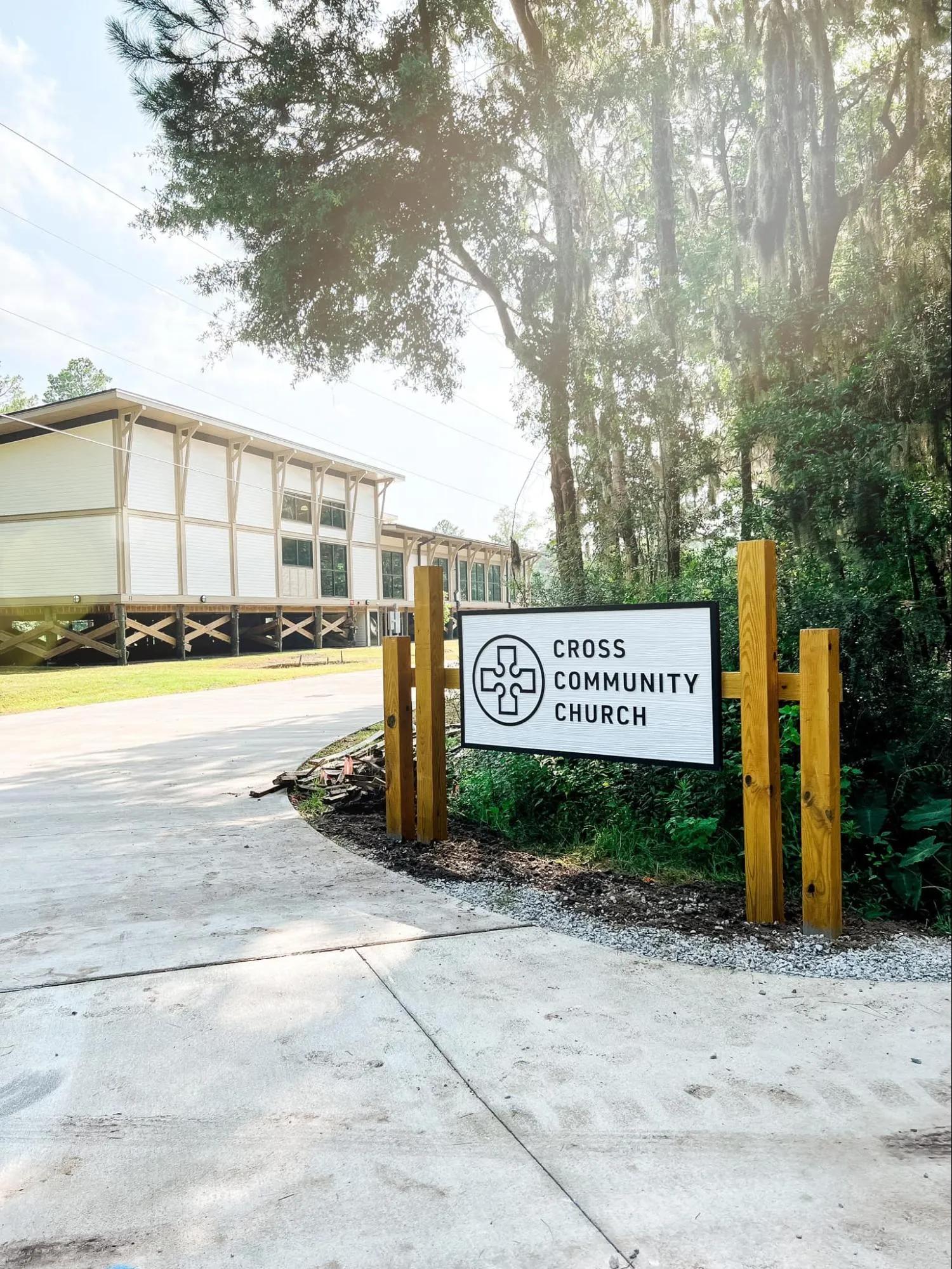 A sign for Cross Community Church stands near a driveway, with a building and trees in the background.