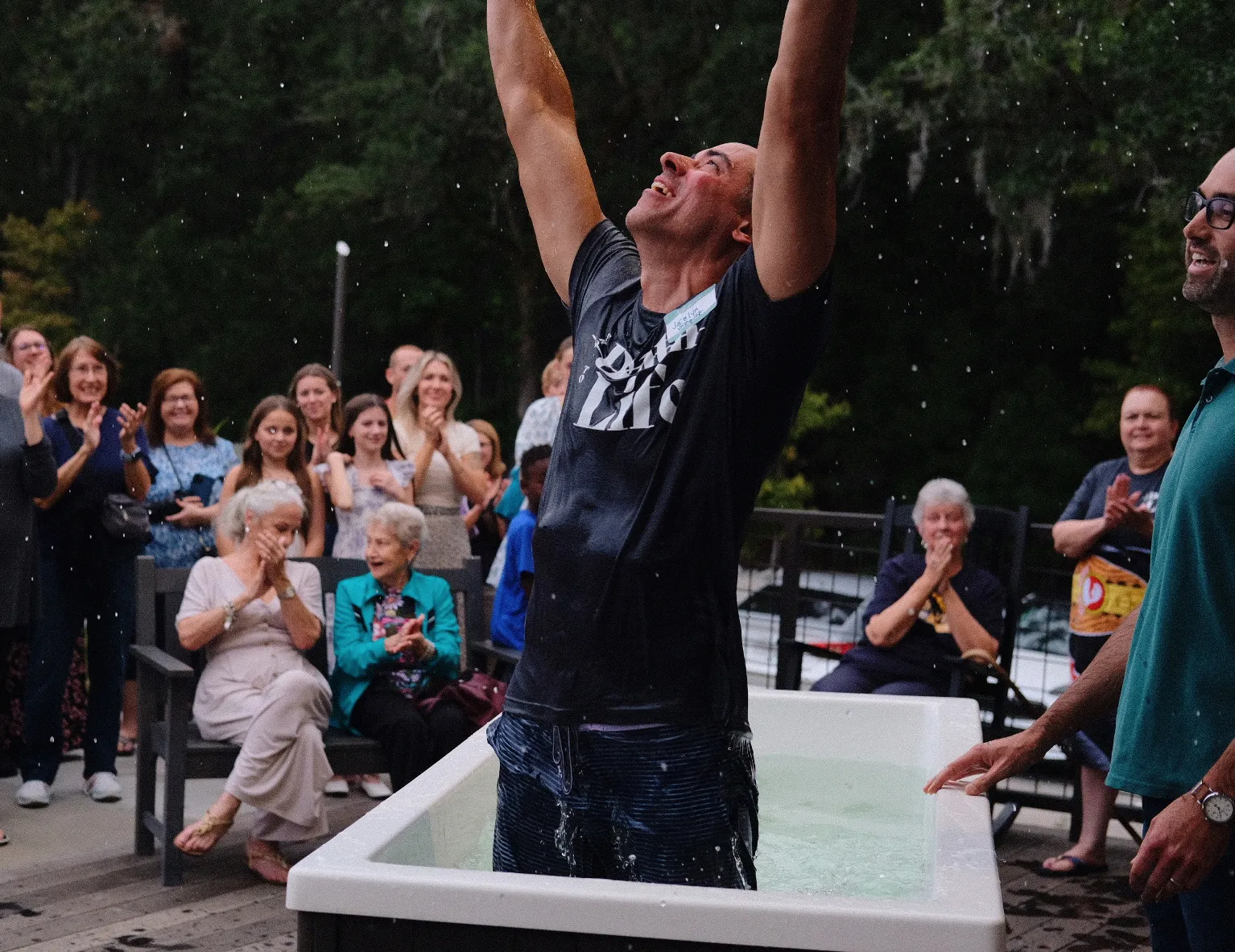 A man celebrates in a baptism pool as a crowd of people applaud and smile outdoors.