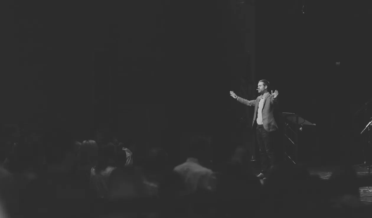 A person stands on stage with arms open, facing an audience in a dark auditorium.
