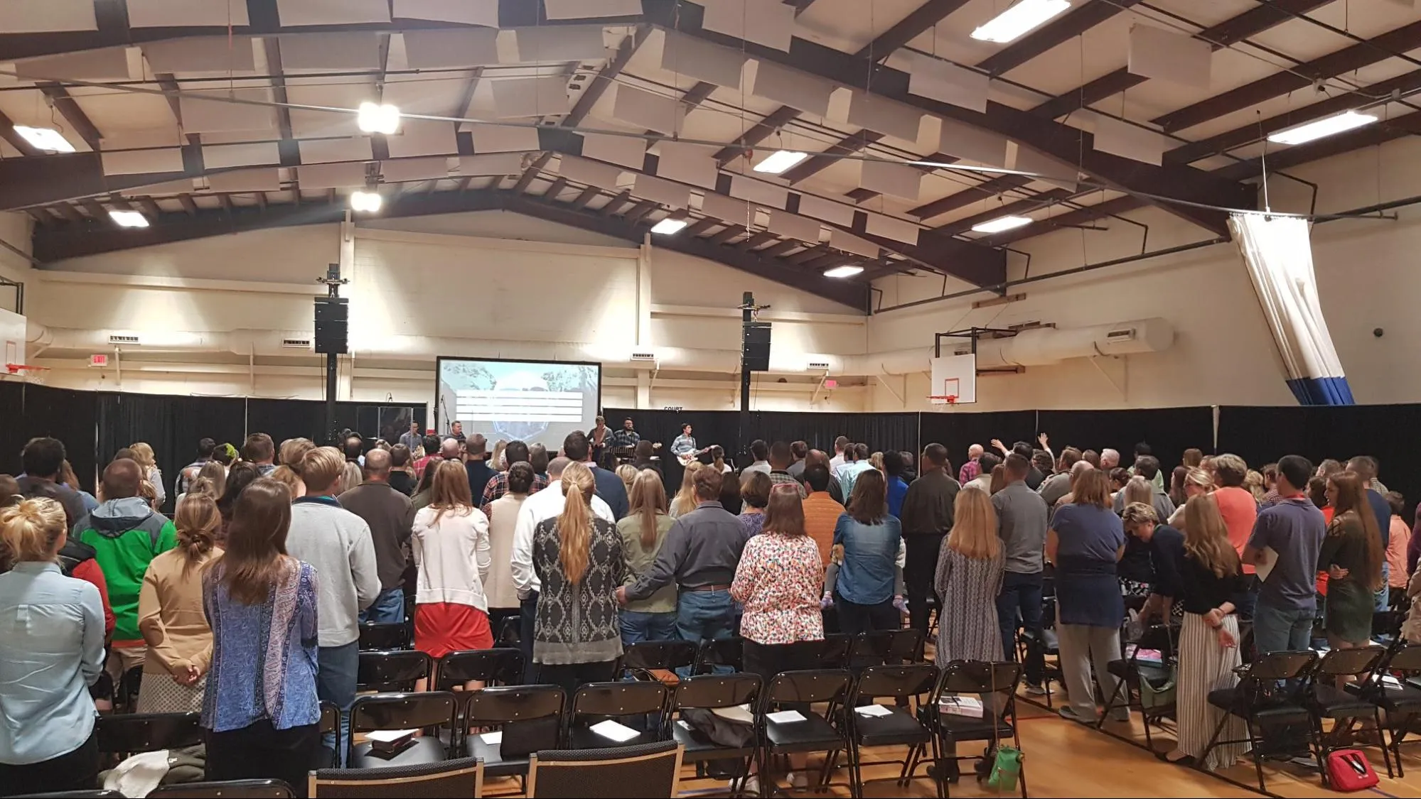 A large group of people stand and face a stage with a screen in a gymnasium during an event or service.