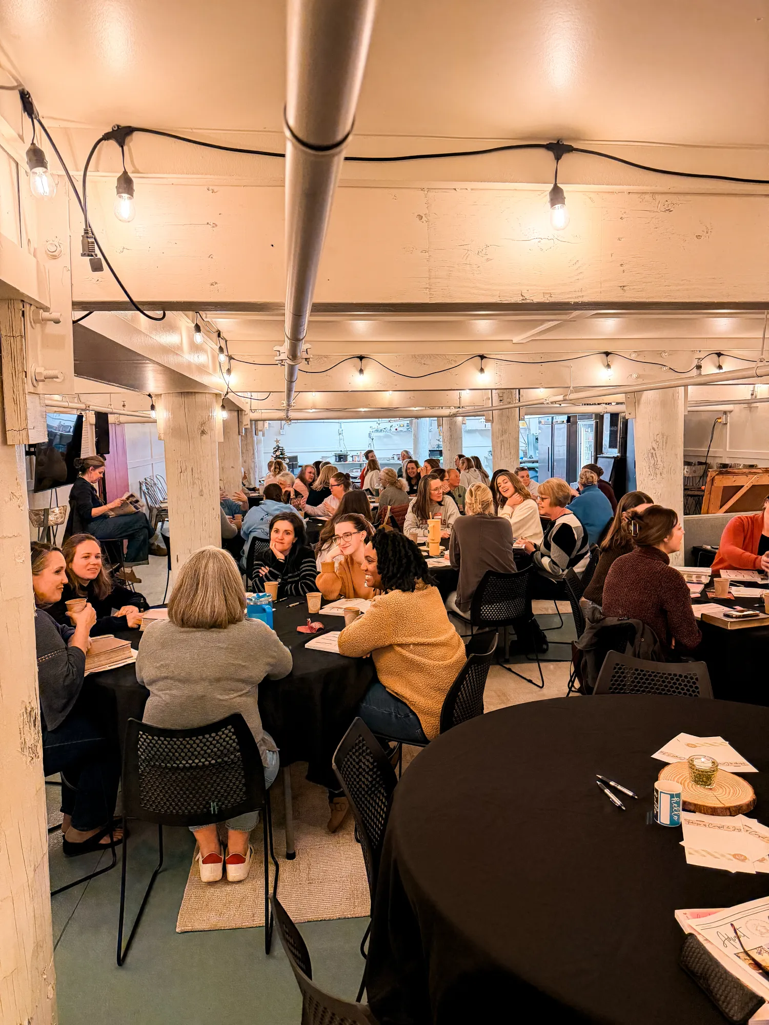 People sit around tables in a warmly lit room, engaged in conversation and eating together.