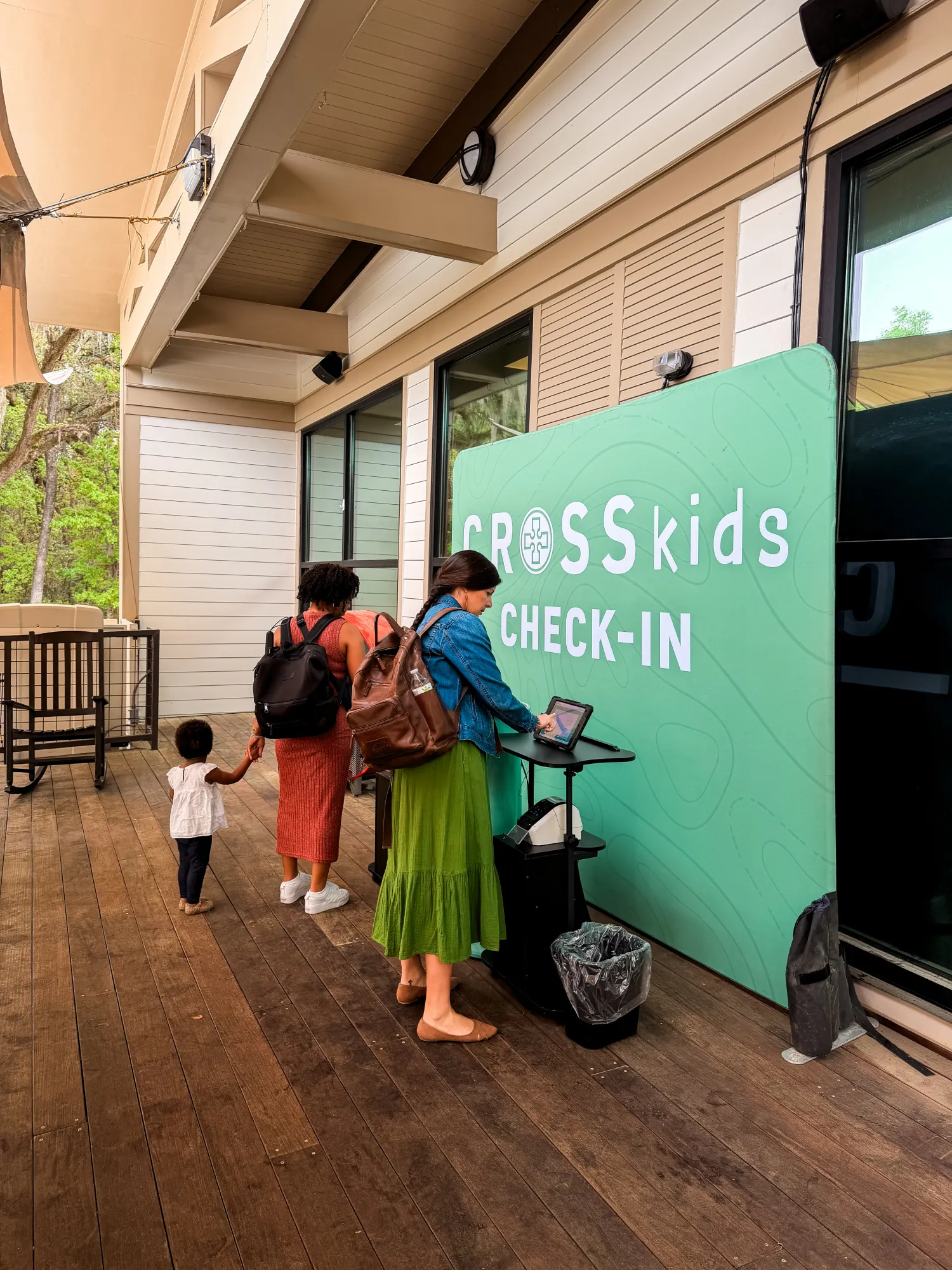 Two women and a child check in at a "CROSS Kids Check-In" station inside a modern building.
