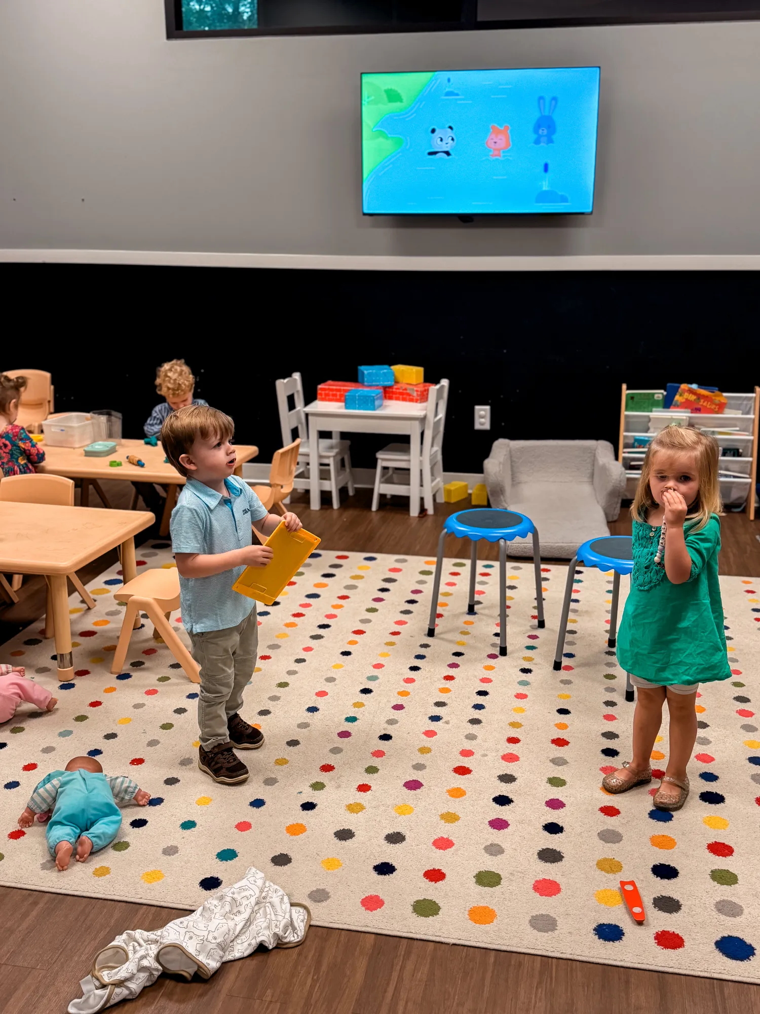Four young children play in a colorful classroom with toys, tables, and a cartoon on a TV screen.
