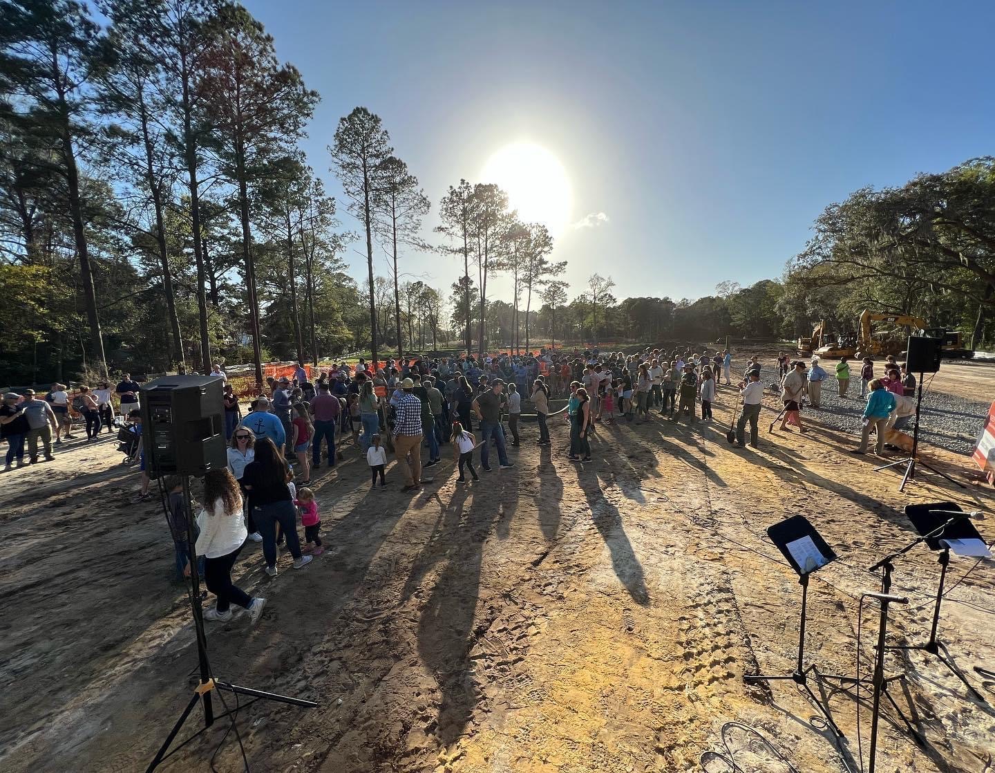 A large crowd gathers outdoors on a dirt lot under tall trees with the sun low in the sky.