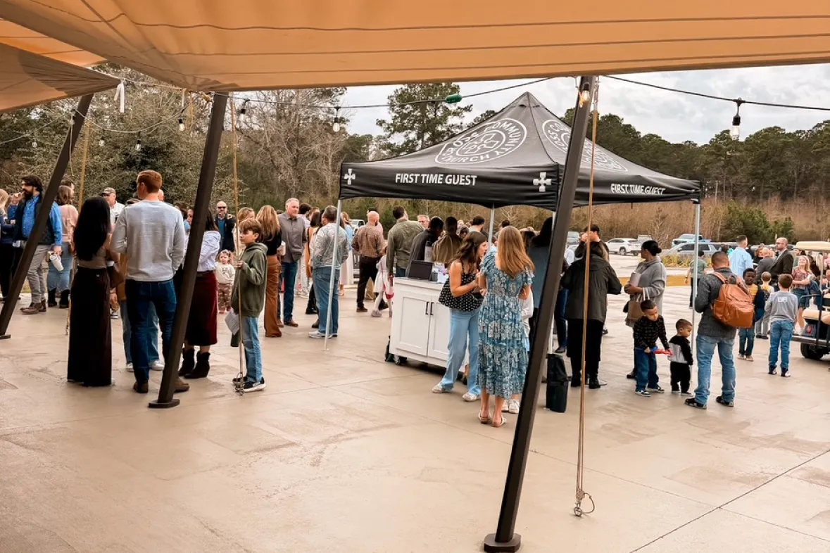 NextStepsCard 1 People gather outdoors under a tent labeled “First Time Guest” on a cloudy day, with trees in the background.