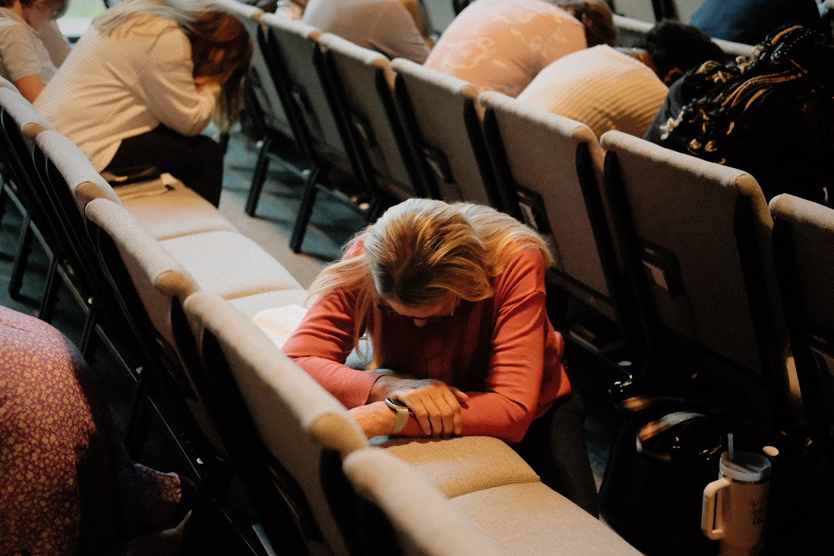 Edited in Tezza with: Wildflower, Brightness, Contrast, Vignette, & Highlights A woman kneels and prays on a church pew, with others also bowing their heads in prayer around her.
